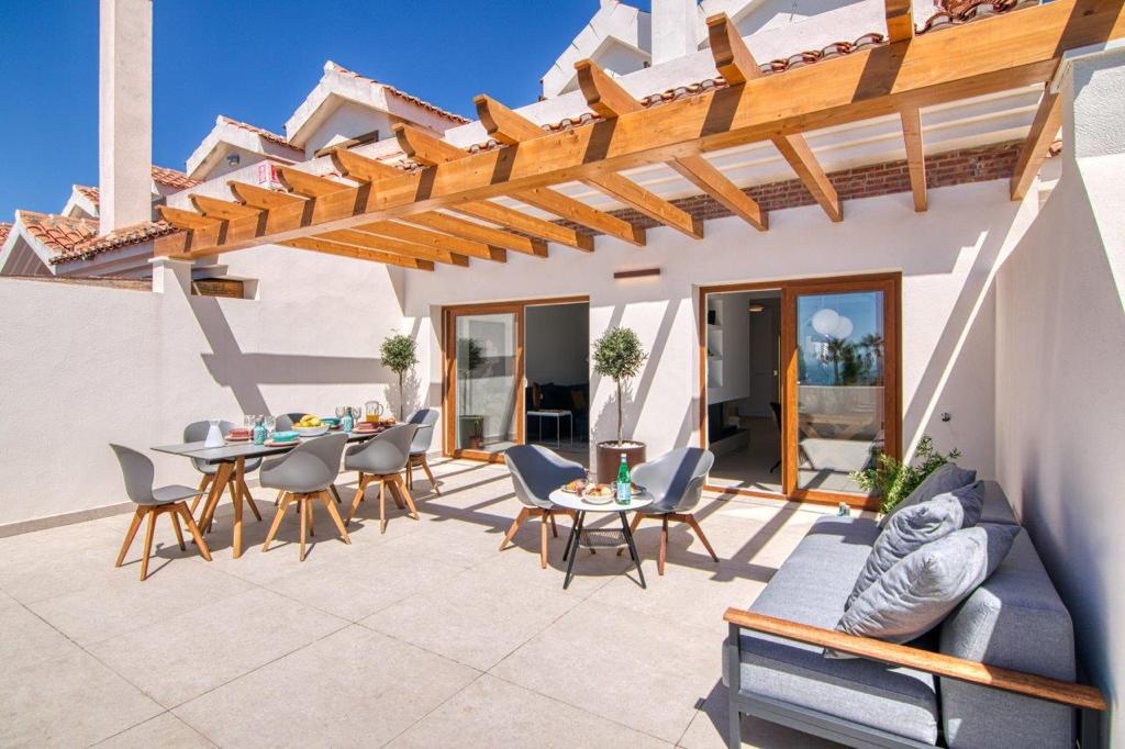 A spacious, contemporary outdoor terrace with light-colored tile flooring sits beneath a wooden-beamed pergola. The outdoor area features a large dining table with gray chairs, a central coffee table with two armchairs, and a sofa with gray cushions to the right, all connected to the interior of the house through large sliding glass doors with wooden frames.