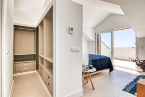 A modern master bedroom with white sloping ceilings. To the left, an open dressing area with light wood wardrobes and drawers stands out, separated by a partition from the sleeping area, where there is a bed with a dark blue bedspread, a round bedside table, and a large sliding glass door that opens onto a sunny terrace.