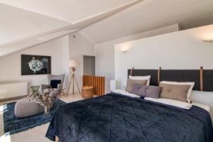 A spacious, contemporary bedroom beneath white, sloping ceilings. In the foreground, a bed dressed with a dark blue quilted bedspread and gray cushions stands out against a dark headboard secured by leather straps. To the left, a reading nook features a gray wingback chair, a pouf on a blue rug, a wooden tripod floor lamp, and a large dark painting on the wall.