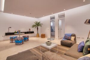 Un amplio salón de diseño contemporáneo y techos bajos con iluminación empotrada. En primer plano destaca un sofá modular marrón con cojines decorativos azules y verdes sobre una alfombra de rayas, junto a una mesa de centro cuadrada blanca y un sillón a juego con una manta azul. Al fondo a la izquierda se sitúa una zona de juegos o reunión con varios pufs de estampado geométrico azul y marrón alrededor de una pequeña mesa blanca, complementado por una palmera decorativa en la esquina y una puerta abierta hacia el pasillo.