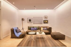 Un salón de diseño moderno con un sofá modular marrón y un sillón a juego decorados con cojines azules y verdes junto a una manta azul. En el centro destacan dos mesas de centro, una redonda blanca y otra cuadrada, dispuestas sobre una alfombra de rayas marrones, mientras que la pared blanca del fondo exhibe una composición de cuadros abstractos, un mapamundi dorado y un adorno en forma de sol bajo un techo con iluminación perimetral.