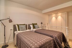 A modern double bedroom with two single beds dressed in three-dimensional geometric patterned bedspreads in brown tones and matching decorative cushions. To the left, a metallic black flexible floor lamp stands out, while in the background, a row of built-in white wardrobes illuminated by warm ceiling spotlights is visible next to a white railing.