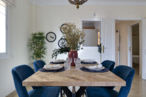 Un comedor moderno con una mesa de madera vestida con platos y copas, rodeada por sillas de terciopelo azul. En el centro destaca un jarrón granate con plantas silvestres secas. Al fondo se aprecian paredes blancas con platos decorativos colgados, una planta alta y dos puertas de paso abiertas hacia el pasillo.
