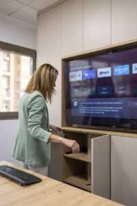 Esperanza con chaqueta verde interactúa con un mueble de oficina integrado de madera clara, el cual aloja un televisor inteligente de gran tamaño que muestra un menú de aplicaciones en pantalla. En primer plano se aprecia una mesa de madera con un teclado de ordenador.
