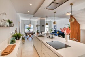 A bright and open-plan space with a contemporary design seamlessly integrates the kitchen, dining room, and living room. A white kitchen island with an integrated induction cooktop and a vase of decorative coral branches takes center stage in the foreground, flanked on the left by a low countertop displaying plants and kitchen accessories. In the background, a wooden dining table sits beneath pendant lights, and a seating area features a dark blue sofa next to a wood-clad staircase on the right.