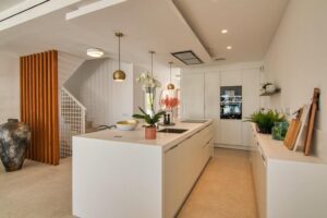 A minimalist kitchen with a large white central island that integrates the sink, decorated with a pot of orchids and a gold bowl beneath spherical bronze pendant lights. To the right, a low linear unit with wooden cutting boards and plants connects to columns of white built-in cabinets at the back with integrated appliances, while to the left, a wooden slatted structure stands out next to a staircase.
