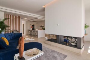 A modern, open-plan living and dining room is divided by a three-sided fireplace set into a large central white column. In the foreground on the left, a curved sofa upholstered in dark blue velvet with mustard cushions sits on a gray rug, next to a side table with decorative items. In the background, the minimalist white kitchen opens up, featuring a central island and several bar stools, flanked by a staircase with decorative wooden slats.