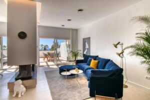 A contemporary living room is dominated by a dark blue velvet sofa with mustard cushions, positioned next to a round pouf and a coffee table on a gray rug. To the left, a freestanding fireplace integrated into a white column with a black clock stands out, while at the far end, a large sliding glass door opens onto a sunny terrace with an outdoor dining table and sea views.