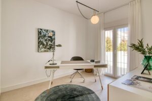 A minimalist office or study area features a white desk with sleek lines and legs with wooden details, accompanied by a black office chair. A dark green round rug takes center stage, illuminated by a pendant lamp with a white glass sphere, while in the background, a double-leaf glass door with white curtains leads to the outside, complemented by an artistic painting in shades of green on the wall and a classic flexible arm lamp