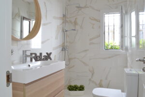 Un cuarto de baño moderno con paredes revestidas de azulejos con efecto mármol calacatta. A la izquierda destaca un mueble de lavabo suspendido de madera clara con un espejo redondo encima y la palabra decorativa "Relax" sobre el grifo. Al fondo se ubica la zona de ducha con grifería cromada y una pequeña ventana abierta que aporta luz natural.