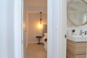 Vista desde el pasillo que muestra a la derecha parte de un cuarto de baño con mueble de lavabo de madera y espejo redondo. Al fondo, a través de una puerta abierta, se vislumbra un dormitorio con una cama vestida de blanco, una mesita de noche redonda oscura y una pared decorada con papel pintado de rayas verticales iluminada por una lámpara colgante.