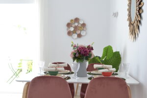 Un comedor moderno centrado en una mesa blanca vestida con vajilla y copas de cristal tallado junto a sillas de terciopelo rosa. En el centro destaca un jarrón gris con flores en tonos malva, mientras que la pared del fondo cuenta con un adorno floral metálico y un espejo de sol dorado a la derecha.