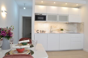 Una cocina americana de diseño moderno con muebles blancos de líneas limpias y electrodomésticos integrados bajo una iluminación empotrada cálida. Al frente destaca un salpicadero con efecto mármol y letras decorativas, conectando en primer plano a la izquierda con una mesa de comedor vestida con platos, copas y un jarrón de flores malvas junto a una silla rosa.