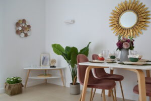 Un comedor de diseño contemporáneo con una mesa blanca vestida con vajilla y copas al lado de sillas de terciopelo rosa. Al fondo destaca una planta de hojas grandes en una maceta gris junto a una mesa auxiliar triangular blanca y madera, decorada con paredes blancas que lucen un espejo de sol dorado y un adorno floral metálico.