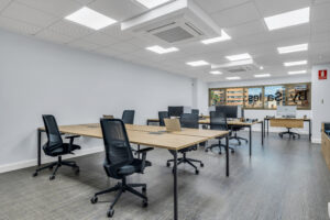 Una amplia sala de trabajo con escritorios de madera y sillas de oficina negras dispuestas en filas paralelas. Al fondo se sitúa un gran ventanal con vinilos corporativos y en el techo destaca una instalación de paneles de iluminación LED.