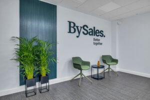 Una zona de espera corporativa con dos sillones giratorios verde oliva y una pequeña mesa auxiliar redonda en el centro. Al fondo, la pared blanca muestra el logotipo "BySales. Better together" junto a un panel vertical de listones azul oscuro y dos plantas altas en maceteros negros.