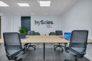 Un gran escritorio de madera clara con ordenadores portátiles abiertos y sillas de oficina ergonómicas negras en primer plano. Al fondo se observa el logotipo corporativo "BySales. Better together" en una pared blanca, junto a un panel de listones oscuros y plantas.