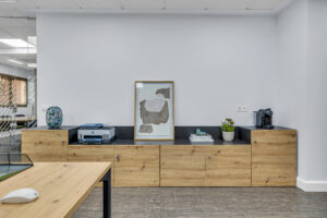 Primer plano de un mueble aparador bajo de madera clara y encimera negra que sostiene una impresora, un cuadro abstracto en tonos grises, libros, plantas y una cafetera de cápsulas. A la izquierda se vislumbra el cristal de una sala de reuniones