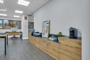 Un mueble aparador bajo de madera clara y encimera negra con una cafetera de cápsulas, libros, plantas decorativas y un cuadro apoyado en la pared. Al fondo se abre la zona de oficinas con escritorios y un ventanal exterior.