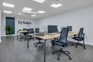 Una zona de trabajo de planta abierta con mesas de madera clara dispuestas en paralelo y sillas de oficina ergonómicas negras. Al fondo, una pared blanca muestra el logotipo corporativo "BySales", flanqueada por un panel decorativo de listones azul oscuro y dos plantas en maceteros negros.