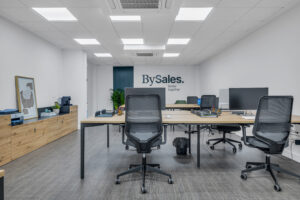 Una amplia zona de trabajo común con escritorios alargados de madera clara y sillas ergonómicas negras. Al fondo, la pared blanca luce el logotipo corporativo "BySales. Better together", junto a un mueble aparador bajo de madera a la izquierda y un falso techo con paneles LED.