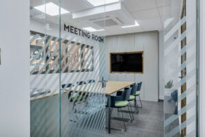 Vista a través de una mampara de cristal con vinilos decorativos de líneas diagonales y el texto "MEETING ROOM". En el interior se aprecia una sala de reuniones con mesa de madera, sillas negras con cojines verdes y una pantalla integrada en un mueble gris al fondo.