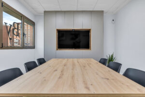 Una larga mesa de reuniones de madera clara en primer plano, rodeada de sillas de oficina negras. Al fondo, un gran televisor integrado en un armario modular gris de suelo a techo, junto a una ventana a la izquierda y una planta verde en la esquina.