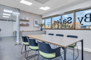 Una sala de reuniones moderna con una gran mesa de madera clara rodeada de sillas oscuras con cojines verdes. Al fondo se observa un ventanal con vinilos corporativos invertidos y, a la izquierda, una mampara de cristal que da acceso a otra sala contigua.
