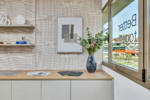Un mueble de oficina bajo en color beige con superficie de madera clara sostiene folletos corporativos y un jarrón de vidrio azul con flores junto a un gran ventanal. En la pared del fondo destaca un cuadro abstracto gris y dos estanterías flotantes con objetos decorativos.