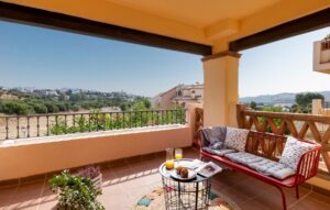Una terraza cubierta de estilo mediterráneo con suelo de baldosas de barro y un banco de forja rojo con cojines estampados. En el centro destaca una mesa auxiliar redonda de metal negro con un aperitivo, con vistas despejadas a un entorno rural y edificaciones blancas bajo un cielo despejado.